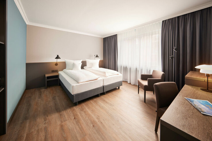 Helles Hotelzimmer mit Doppelbett, Schreibtisch, zwei Sesseln und großen Fenstern mit Vorhängen.