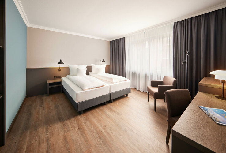 Helles Hotelzimmer mit Doppelbett, Schreibtisch, zwei Sesseln und großen Fenstern mit Vorhängen.