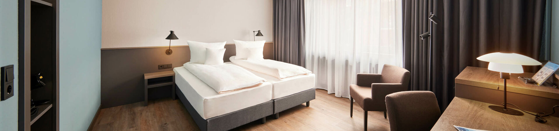 Comfort Plus Zimmer im ATLANTIC Hotel Landgut Horn Helles Hotelzimmer mit Doppelbett, Schreibtisch, zwei Sesseln und großen Fenstern mit Vorhängen.