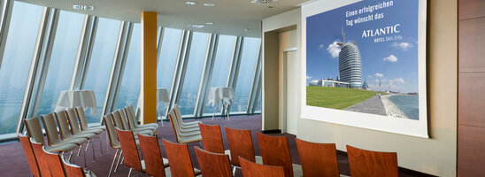 Moderner Konferenzraum im ATLANTIC Hotel Sail City mit Reihenstühlen und Blick auf eine Präsentationsleinwand.