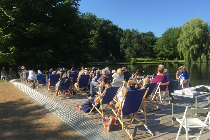 Menschen entspannen in Liegestühlen auf einer Terrasse am See, umgeben von grünen Bäumen unter blauem Himmel.