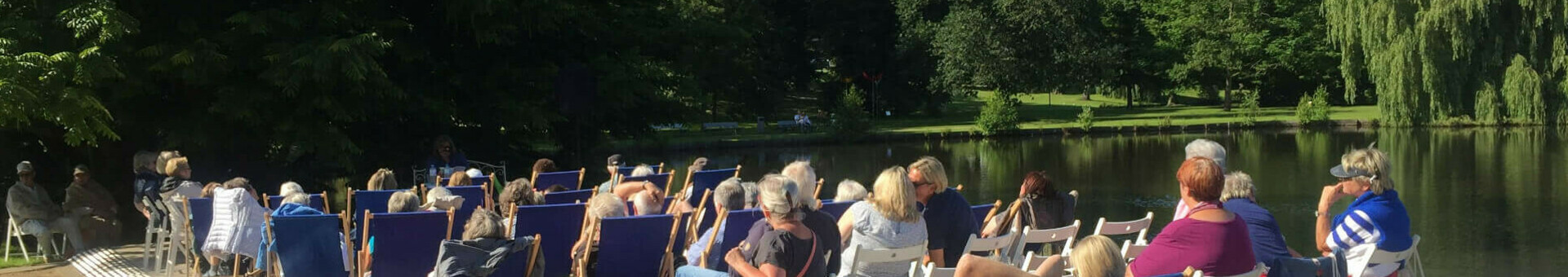Menschen entspannen in Liegestühlen auf einer Terrasse am See, umgeben von grünen Bäumen unter blauem Himmel.