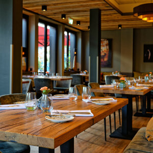 Gemütliches Restaurant mit Holztischen, gedeckt mit Gläsern und Tellern, warme Beleuchtung, große Fenster.