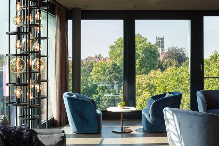 Cozy lounge with blue armchairs and table, large windows with a view of green trees and church tower in the background.