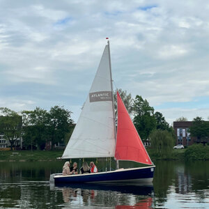 Sailing boat with ATLANTIC Hotel Münster logo, sailing on a calm lake surrounded by green trees.