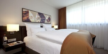 Modern hotel room with double bed, white bed linen, brown bedspread, artwork on the wall and natural light through the window.