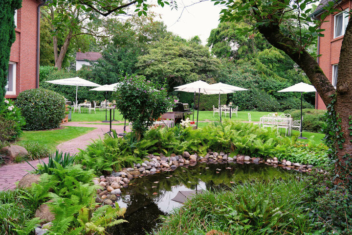 Green garden with pond, white seating areas and parasols at the ATLANTIC Hotel Landgut Horn Bremen.