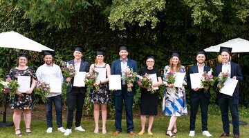 Eine Gruppe von acht Personen in Abschlusskleidung hält Urkunden und Blumensträuße im Garten.