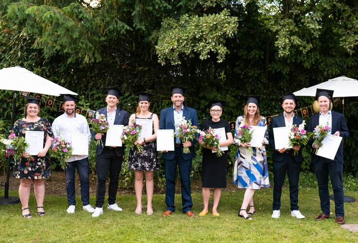 A group of eight people in graduation attire hold certificates and bouquets of flowers in the garden.