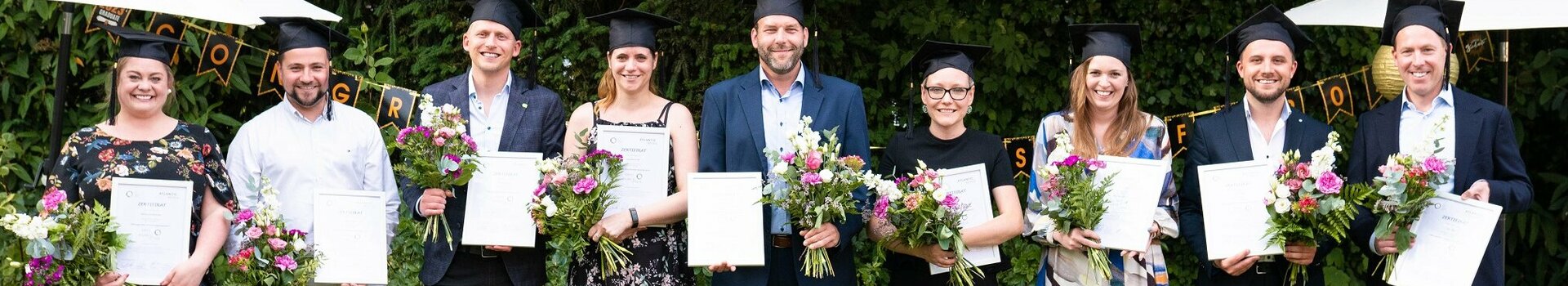 A group of eight people in graduation attire hold certificates and bouquets of flowers in the garden.
