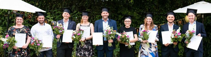 Eine Gruppe von acht Personen in Abschlusskleidung hält Urkunden und Blumensträuße im Garten.
