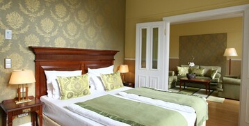 Elegantes Hotelzimmer mit Doppelbett, grüner Bettwäsche, stilvollen Kissen und angrenzendem Wohnbereich mit Sofas.