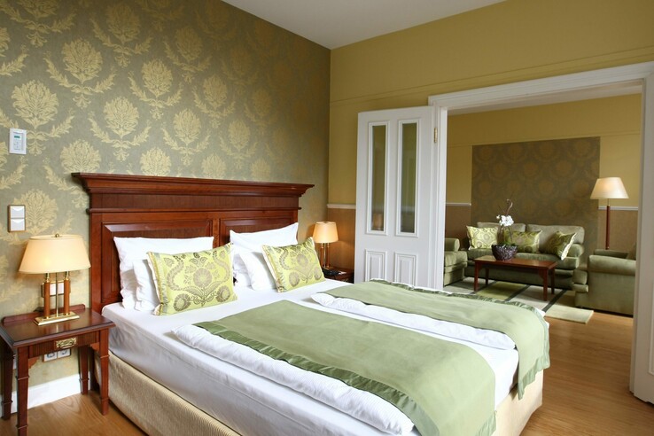 Elegantes Hotelzimmer mit Doppelbett, grüner Bettwäsche, stilvollen Kissen und angrenzendem Wohnbereich mit Sofas.