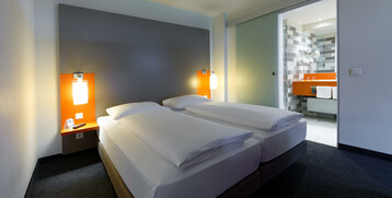 Modern hotel room with two single beds, illuminated headboard and view of the bathroom with orange accents.