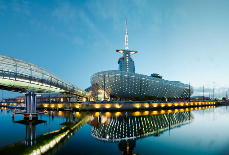 Modernes Gebäude am Wasser mit beleuchteter Fassade und gläsernem Übergang, spiegelnd im ruhigen Wasser bei Dämmerung.