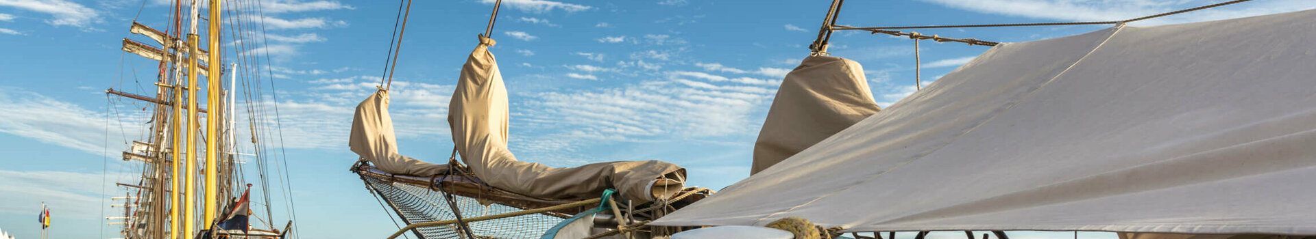 Segelschiffe am Pier unter blauem Himmel mit Wolken, Seile im Vordergrund.
