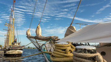 Segelschiffe am Pier unter blauem Himmel mit Wolken, Seile im Vordergrund.