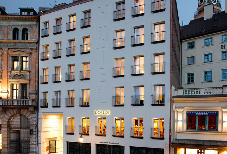 Außenansicht des LOUIS Hotels in München bei Dämmerung, mit beleuchteten Fenstern und umliegenden Gebäuden.