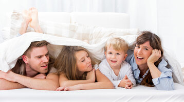 Familie lächelt fröhlich unter einer Bettdecke in einem hellen Hotelzimmer des ATLANTIC Hotel Universum in Bremen.