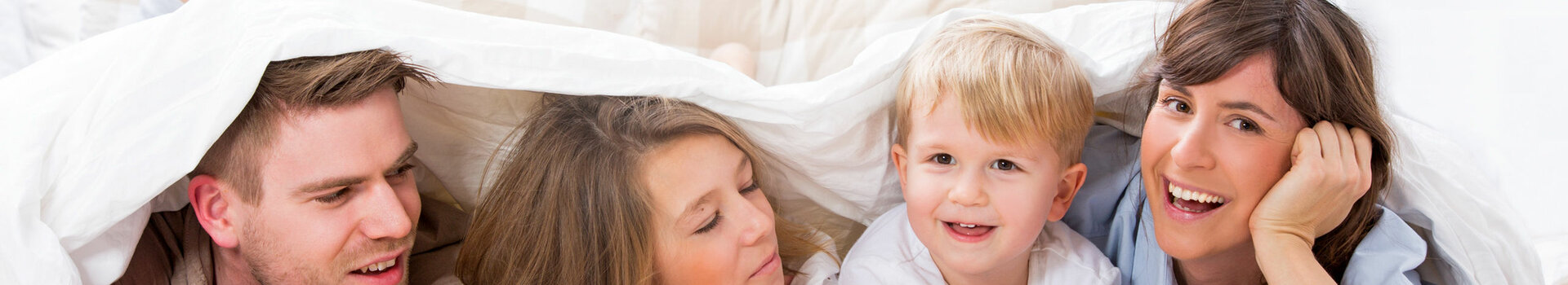 Family smiling happily under a comforter in a bright hotel room at the ATLANTIC Hotel Universum in Bremen.