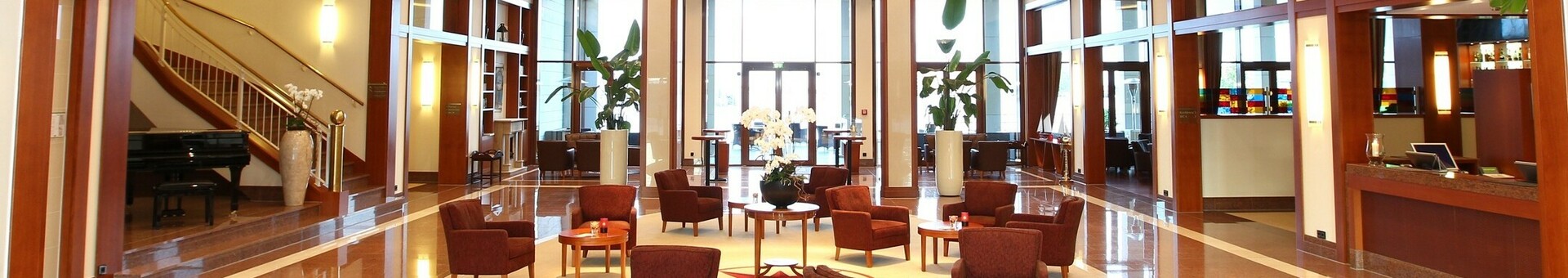 Hotelhalle des ATLANTIC Hotel Wilhelmshaven mit Sitzecke Elegante Hotellobby mit roten Sesseln, runden Tischen, modernen Kronleuchtern und Empfangsbereich rechts.
