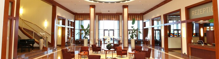 Elegante Hotellobby mit roten Sesseln, runden Tischen, modernen Kronleuchtern und Empfangsbereich rechts.