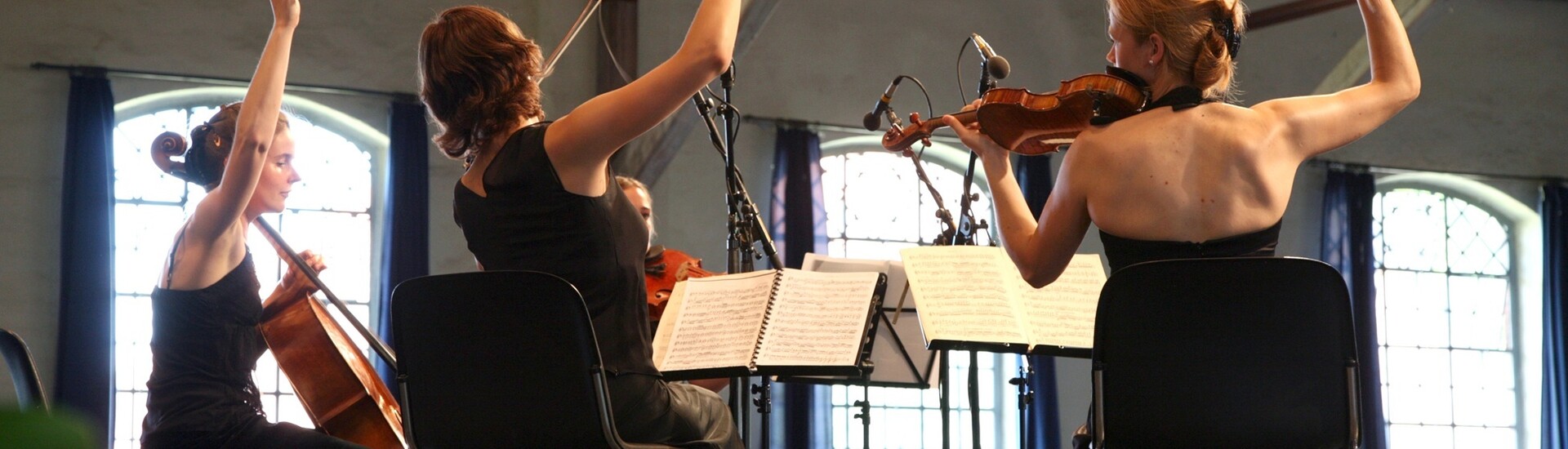 Streichquartett spielt auf Bühne, Musikerinnen in schwarzen Kleidern, Publikum im Hintergrund.
