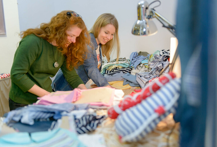 ATLANTIC Hotel Sail City | Upcycling-Offensive "GRÜNER FADEN" Zwei Frauen stöbern lächelnd durch bunte Stoffe auf einem Tisch unter einer Lampe.