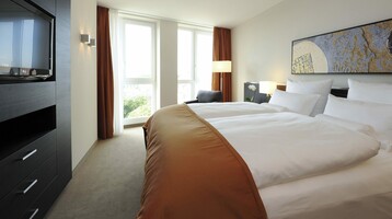 Helles Hotelzimmer mit großem Bett, modernem Dekor, Fenster mit Vorhängen, TV und Kunstwerk an der Wand.