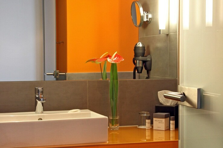 Modernes Badezimmer im ATLANTIC Hotel Kiel mit orangen Wänden, Waschbecken, Spiegel, Föhn und frischen Blumen.