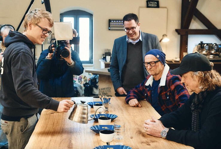 Der Kaffee von Münster Personen bei einer Kaffeeverkostung an einem Holztisch, Fotograf im Hintergrund.