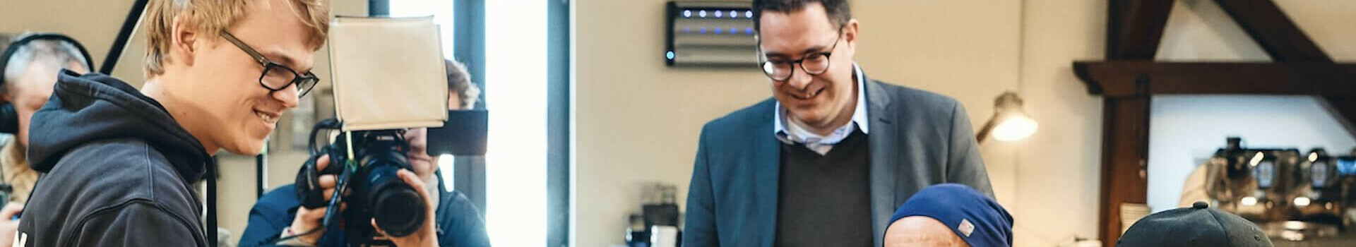 Personen bei einer Kaffeeverkostung an einem Holztisch, Fotograf im Hintergrund.