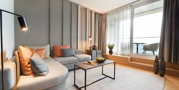 Modern hotel room with gray sofa, colorful cushions, coffee table, lamp and balcony view through large windows.