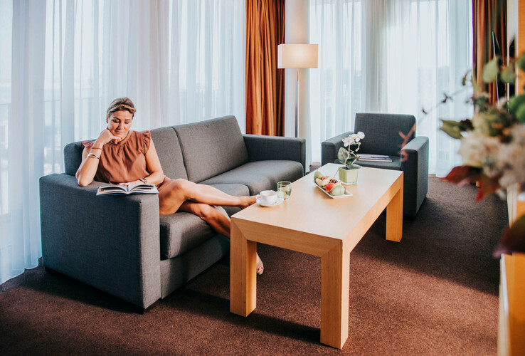 Bright hotel suite with gray sofa, reading lamp, coffee table and woman reading a book.
