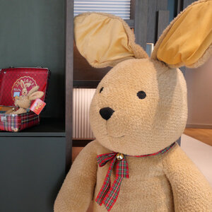 Large stuffed animal with bunny ears and plaid scarf, next to a small suitcase with another stuffed bunny.