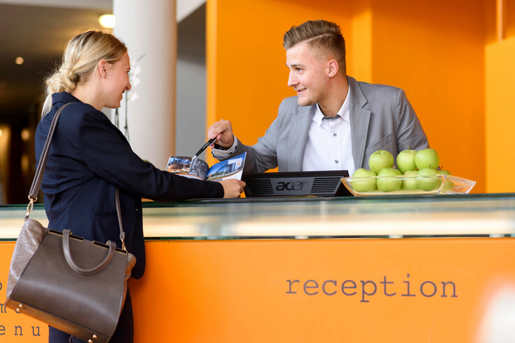 Hotelrezeption mit lächelndem Mitarbeiter, der einer Frau Informationen gibt; Schale mit grünen Äpfeln auf dem Tresen.