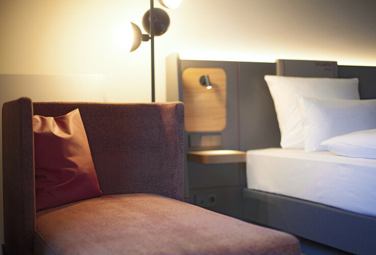 Modern hotel room with cozy armchair, reading lamp and a bed with white pillows at the ATLANTIC Hotel Airport Bremen.