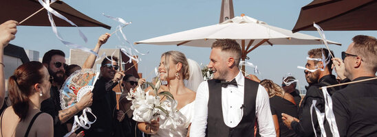 Hochzeitspaar verlässt strahlend eine festliche Zeremonie im Freien, umgeben von jubelnden Gästen mit Bändern.