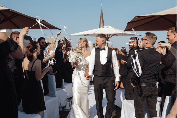 Hochzeitspaar verlässt strahlend eine festliche Zeremonie im Freien, umgeben von jubelnden Gästen mit Bändern.