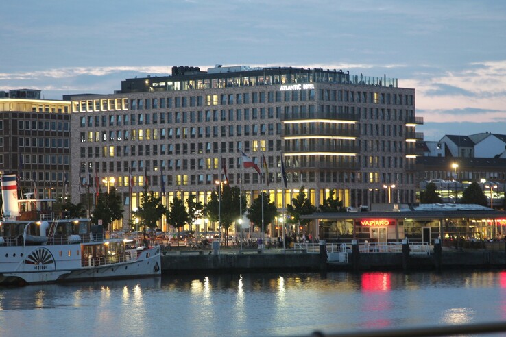 ATLANTIC Hotel Kiel bei Abenddämmerung, beleuchtet, mit Blick auf den Hafen und ein Vapiano-Restaurant am Wasser.