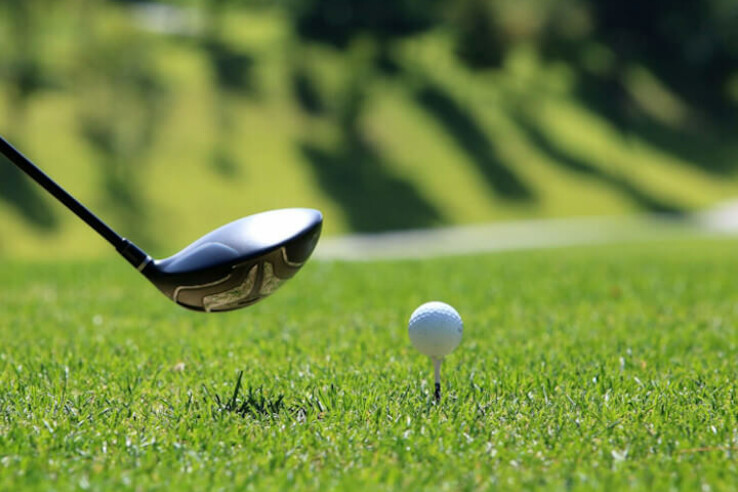 Golf club swinging after golf ball on tee on green grass, with blurred background.