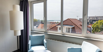 Helles Hotelzimmer mit zwei blauen Sesseln, Tisch, großer Fensterfront und Blick auf Dächer und Bäume.