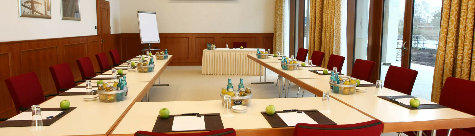 Heller Konferenzraum mit U-förmiger Tischanordnung, roten Stühlen und Tageslicht, ausgestattet für Meetings.