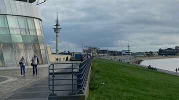 ATLANTIC Hotel Sail City in Bremerhaven, modernes Gebäude am Wasser, Spaziergänger und Radfahrer auf dem Vorplatz.