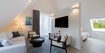 Helles Hotelzimmer mit Sofa, Sesseln, Flachbild-TV, Teppich und Bett im Hintergrund, modernes Design.