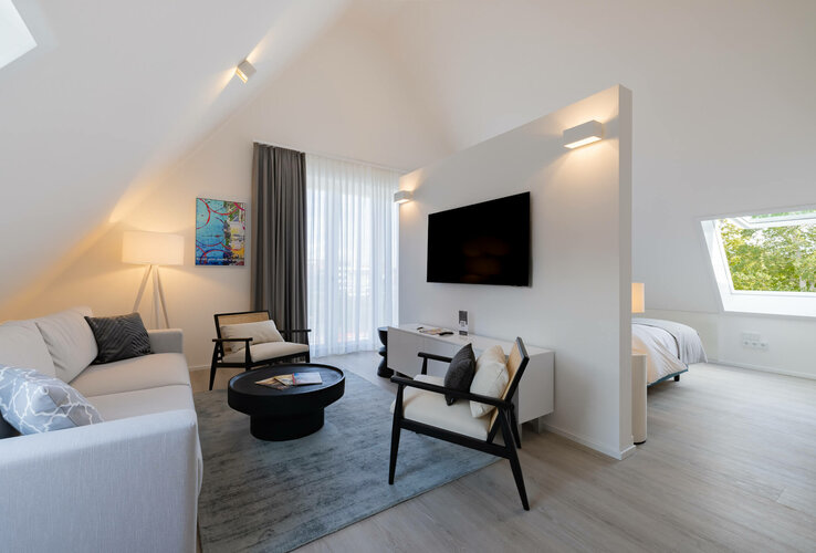 Helles Hotelzimmer mit Sofa, Sesseln, Flachbild-TV, Teppich und Bett im Hintergrund, modernes Design.