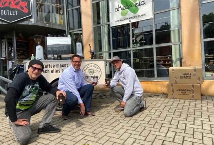 Three men kneel in front of the ATLANTIC Hotel Münster, holding coffee cups, surrounded by signs and cardboard boxes.