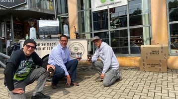 Drei Männer knien vor dem ATLANTIC Hotel Münster, halten Kaffeetassen, umgeben von Schildern und Kartons.