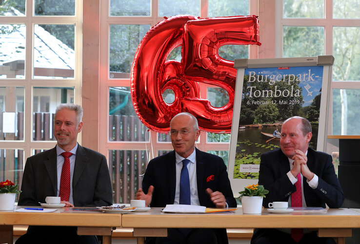 Three men in suits are sitting at a table in front of a window, with a red "65" balloon and a Bürgerpark raffle poster.