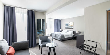 Helles Hotelzimmer mit Doppelbett, Sofa, Tisch, Stuhl und großen Fenstern mit blauen Vorhängen.